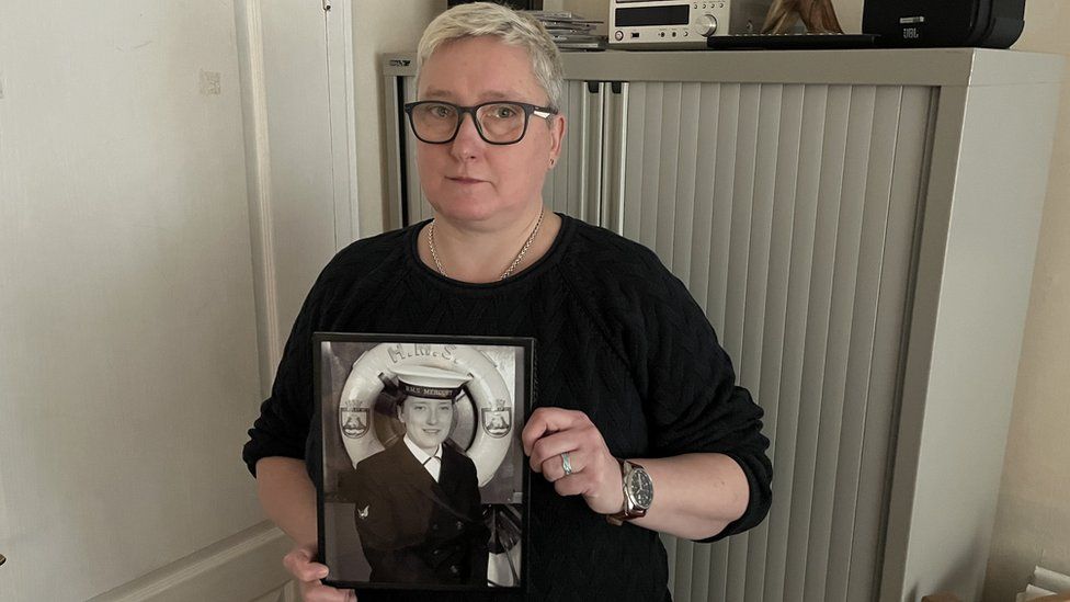 A woman with short blond hair and glasses holds a black and white picture of her younger self in a Navy uniform