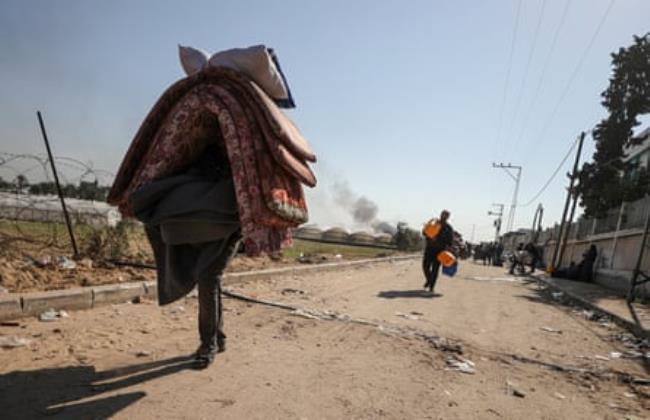 A lone person carries mattresses over the head as they walk down an empty street. Behind them some way away someone else carries bags. In the distance smoke can be seen rising.