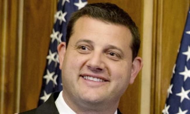 Closeup up young-ish white-presenting man smiling in suit in front of American flag alo<em></em>ngside wood paneling.