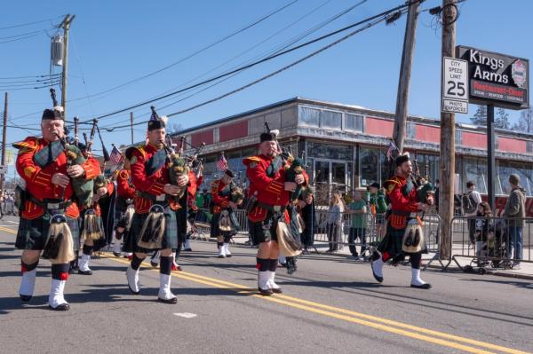 A row of bagpipers in red outfits march down a parade route.