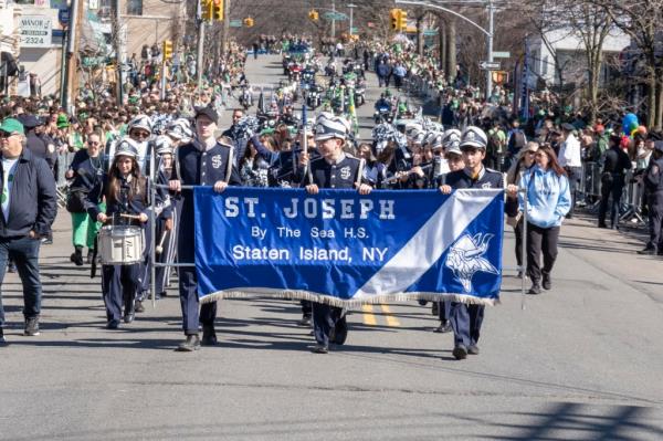 A group of students in band uniforms marching in formation behind a blue banner that says St. Joseph by the Sea HS Staten Island NY