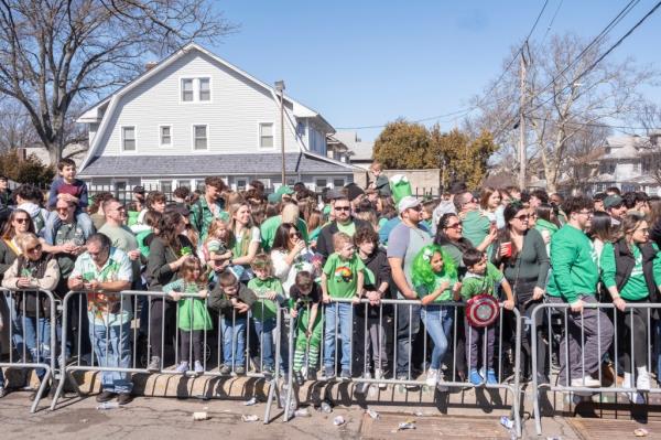 A large group of people on the sidewalk behind a me<em></em>tal barrier in a suburban environment, many of them wearing green.