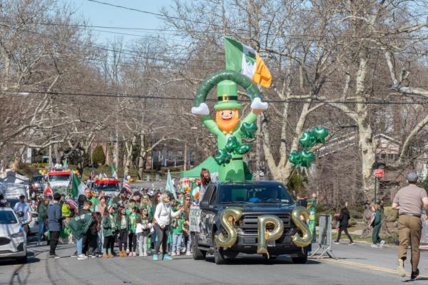 A parade crowd walking behind a truck with a large inflatable leprechaun on top.