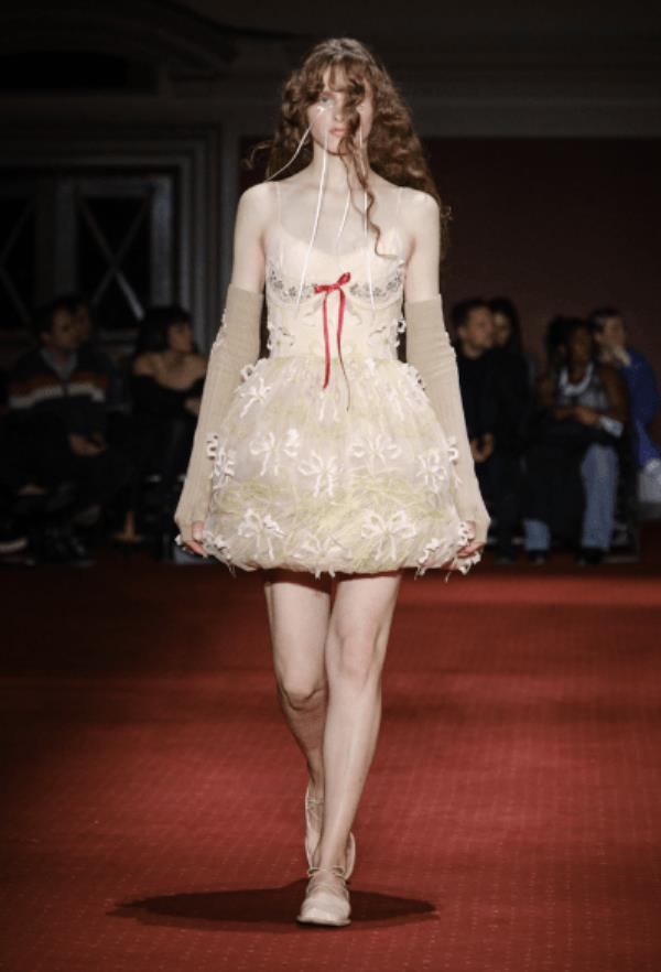 Simone Rocha Model in cream mini dress with structured skirt, adorned with red bow on bodice