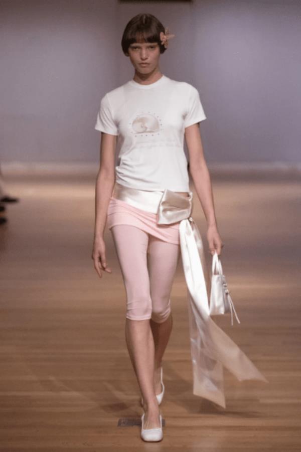 Sandy Liang model wearing white tee and pink knee length shorts with large cream satin bow tied at hip and around waist