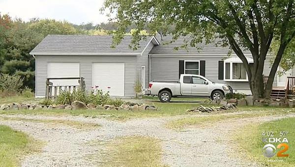 A suburban house with gray siding, black shutters, and two garage doors with a silver pickup truck parked out front and a large tree at the top of a gravel driveway.