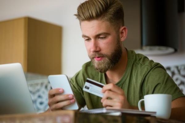 Young bearded man wearing green t-shirt sitting in the kitchen at home, holding smart phone and credit card in hands, shopping online.