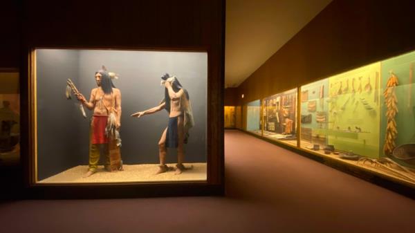 An empty museum with two mannequins in Native American costumes behind glass and to the right glass cases showing Native American objects.