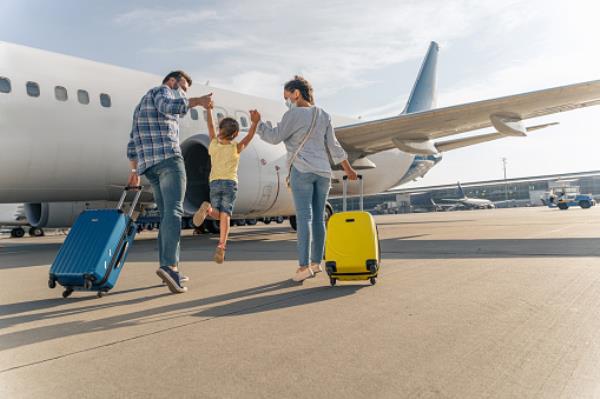 Happy family in masks, walking towards plane. The child is in the middle holding hands with his parents and being held up, as the parents pull suitcases.