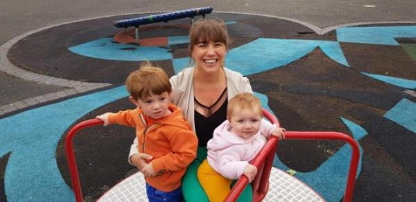 Sarah -on a rounda<em></em>bout in the park with her two kids, looking to camera and smiling