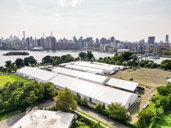 Migrants seeking asylum in US arrive at newly built tents on Randall's Island - large white tents with trees and city in the background.